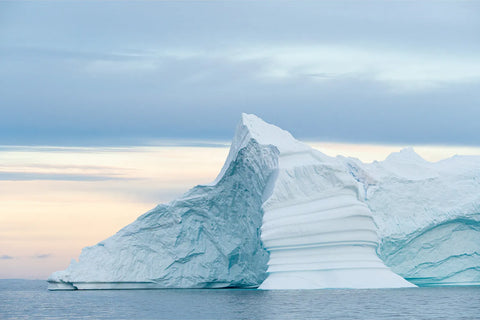 Layered iceberg with horizontal striations rising from still Arctic waters under a soft pastel sky, Greenland