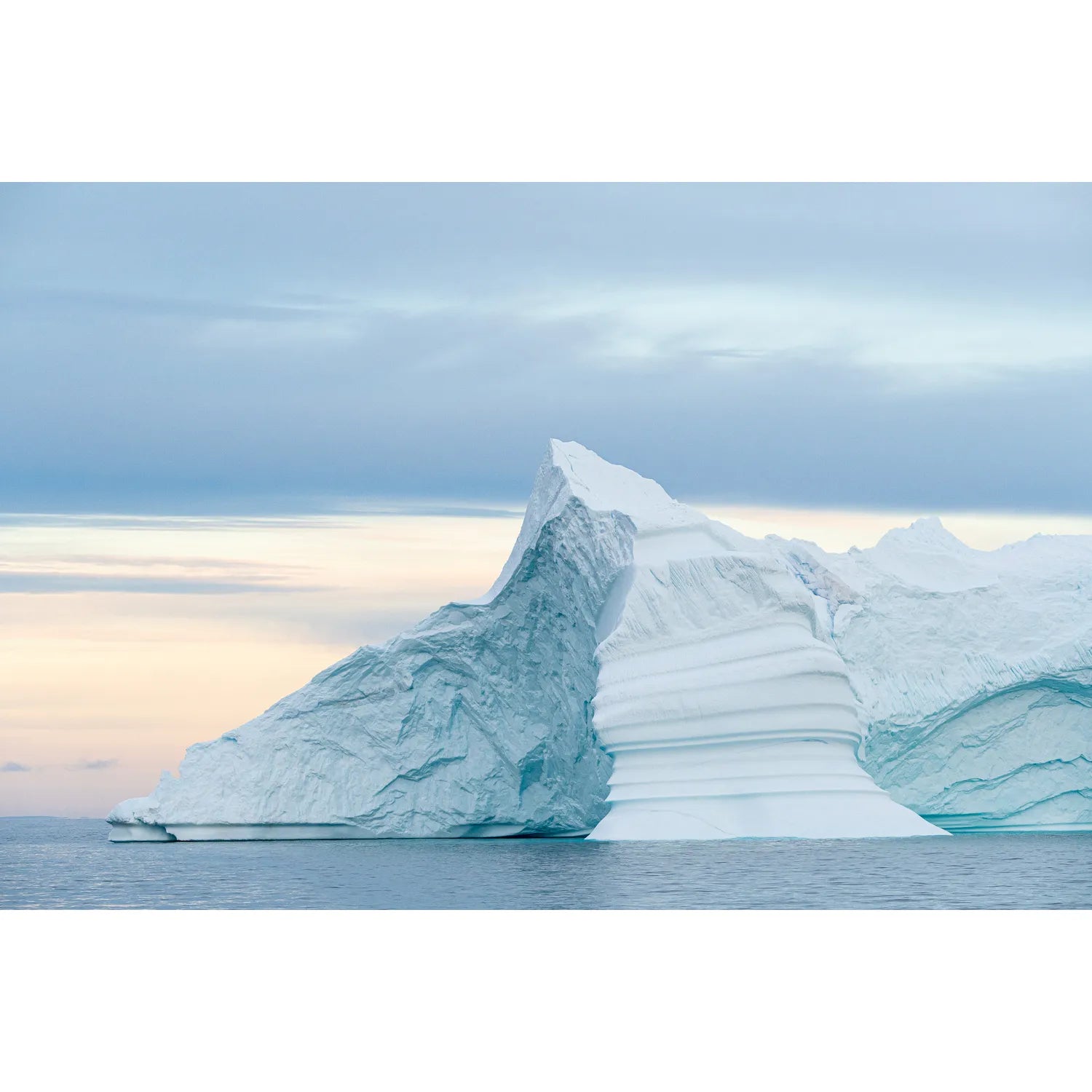 Layered iceberg with horizontal striations rising from still Arctic waters under a soft pastel sky, Greenland