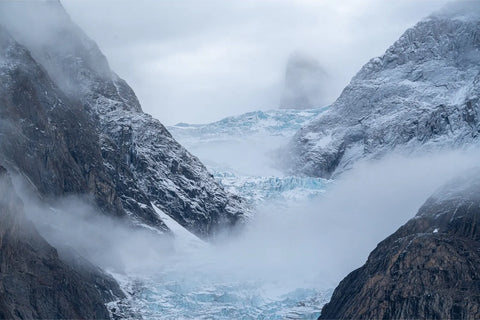 Mist rolling through a glacial valley in Scoresby Sund with blue glacier ice cradled between dark rock faces