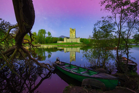 Ross Castle illuminated under purple and blue aurora borealis, reflected in Lough Leane with a green rowing boat and old trees