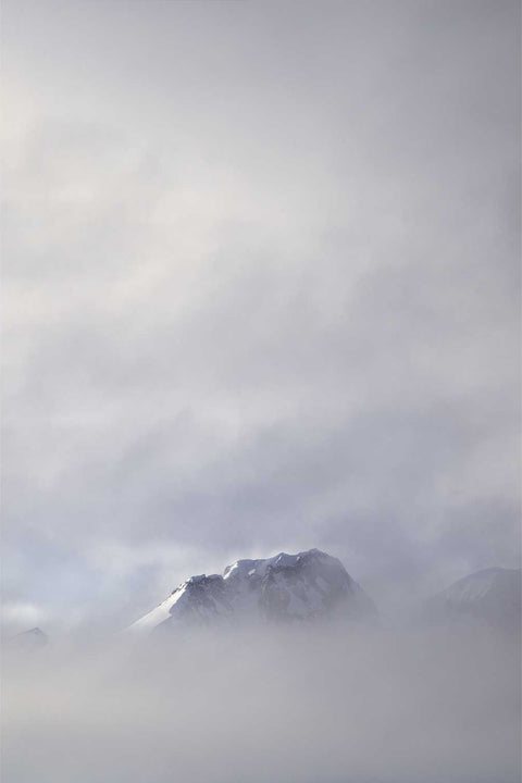 Snow-covered mountain peak emerging from dense low fog above Charlotte Bay on the Antarctic Peninsula in grey monochromatic light