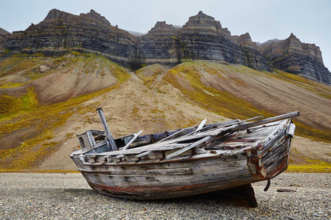 Old silver-grey wooden shipwreck on a gravel beach in Svalbard with dramatically layered mountains rising behind under overcast sky