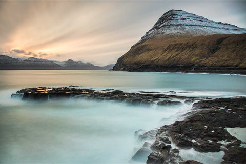 Sunrise over Gjogv village in the Faroe Islands with silky water flowing over dark basalt rocks and snow-capped mountains in the distance, fine art landscape photograph by Peter Cox