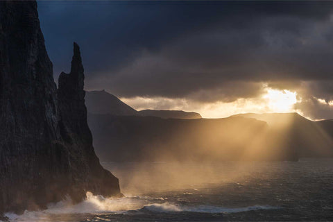 Trollkonufingur sea stack silhouetted at sunrise with golden light breaking through stormy clouds, Faroe Islands