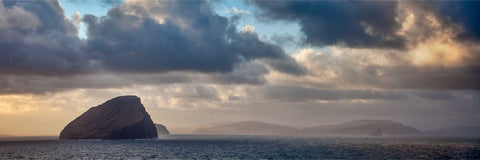 The uninhabited island of Koltur rising from the sea under dramatic clouds with warm light on the horizon, Faroe Islands