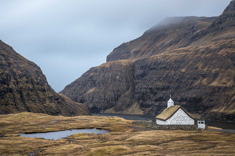 Traditional grass-roofed church in the Saksun valley with towering cliffs, a tranquil lagoon and mountains in the Faroe Islands