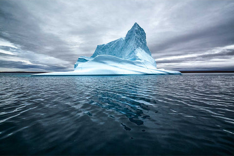 A peaked iceberg glowing blue-white against stormy skies and dark water in Greenland — fine art landscape photograph by Peter Cox