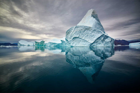 A towering iceberg with perfect reflection in the still waters of Scoresby Sound, Greenland under grey skies — fine art landscape photograph by Peter Cox