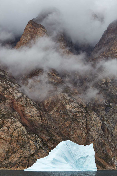 Pale iceberg floating at the base of vast mist-shrouded rock cliffs in Ofjord, Greenland