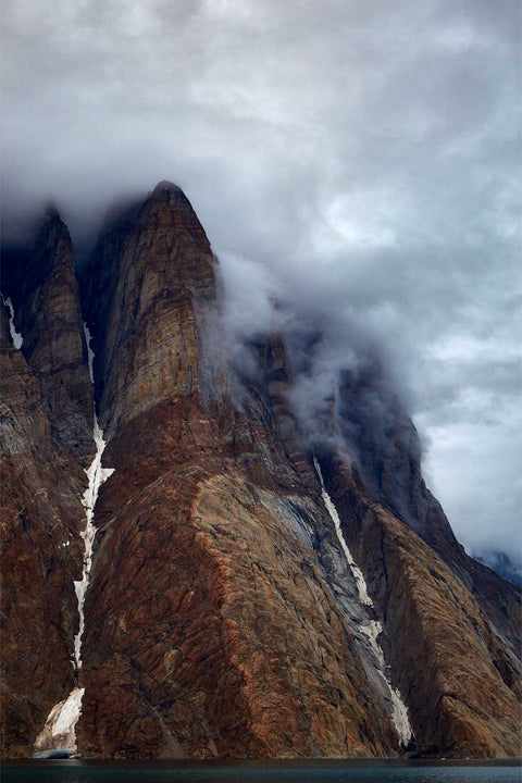 Towering snow-filled cliffs of Ofjord rising dramatically from the water in Greenland, wrapped in cloud