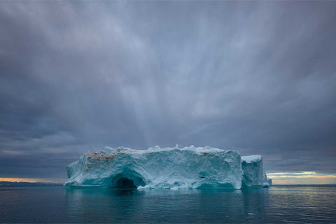 An iceberg glowing in dramatic light against an approaching squall at sunset in Greenland — fine art landscape photograph by Peter Cox