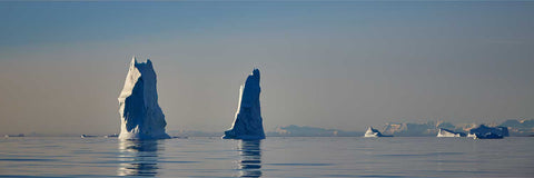 Two towering spire-shaped icebergs rising from glassy calm waters off Greenland under a hazy sky