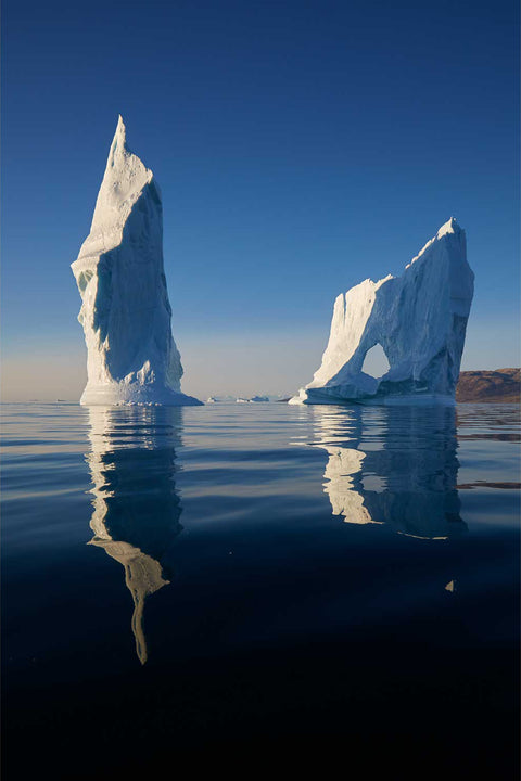 Two tall iceberg towers reflected in still Greenland waters under a pale sky
