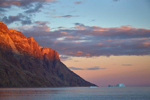 Golden sunset light illuminating rugged Greenland cliffs above calm Arctic waters with icebergs in the distance