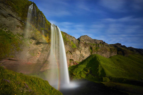 Seljalandsfoss waterfall dropping 60 metres with a rainbow in the mist, green moss-covered cliffs and blue sky in Iceland