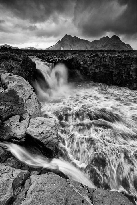 Black and white long exposure of a powerful river cascading through volcanic rock with jagged mountain peaks under stormy skies in Iceland's Fjallabak highlands