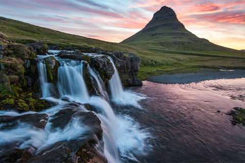 Kirkjufellsfoss waterfall cascading over rocks with Kirkjufell mountain silhouetted against a pink and orange midnight sun sky, Iceland