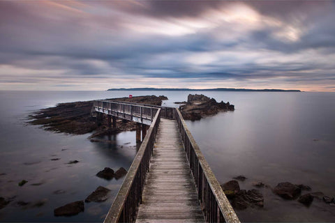 Long exposure of Pan's Rock with its footbridge at dawn on the Antrim Causeway Coast, silky water and blurred clouds, Rathlin Island on the horizon