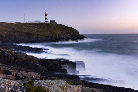 Long exposure of waves crashing against rocks at the Old Head of Kinsale lighthouse at dusk with its lantern shining