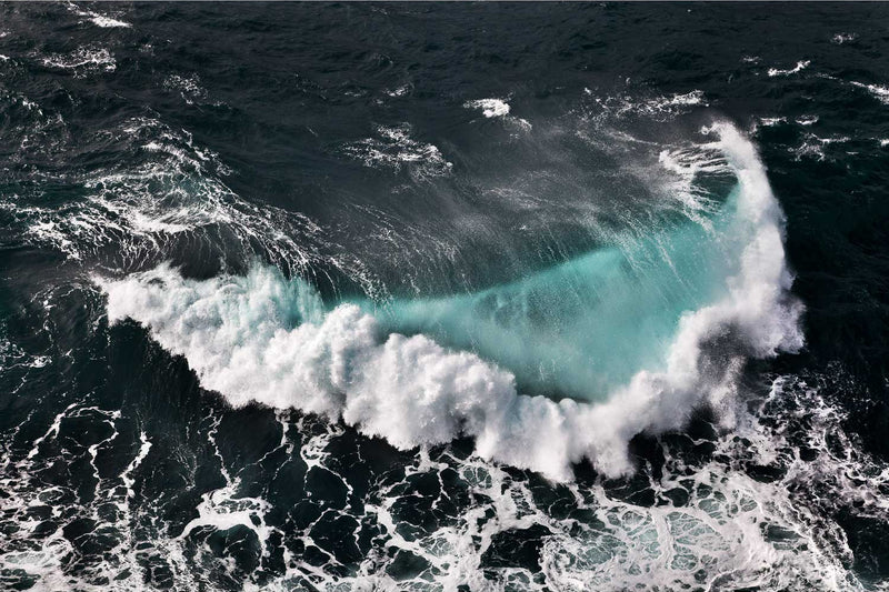 Looking straight down onto near black open water off the Fastnet Rock, Co. Cork, a single wave curls across the frame with light coming through its face as a pane of turquoise. White foam blows off the breaking edge, and older foam laces the darker water below.