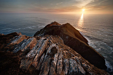 Sunset over Mizen Head signal station and lighthouse at Ireland's most southwesterly point with rocky coastline leading to the setting sun