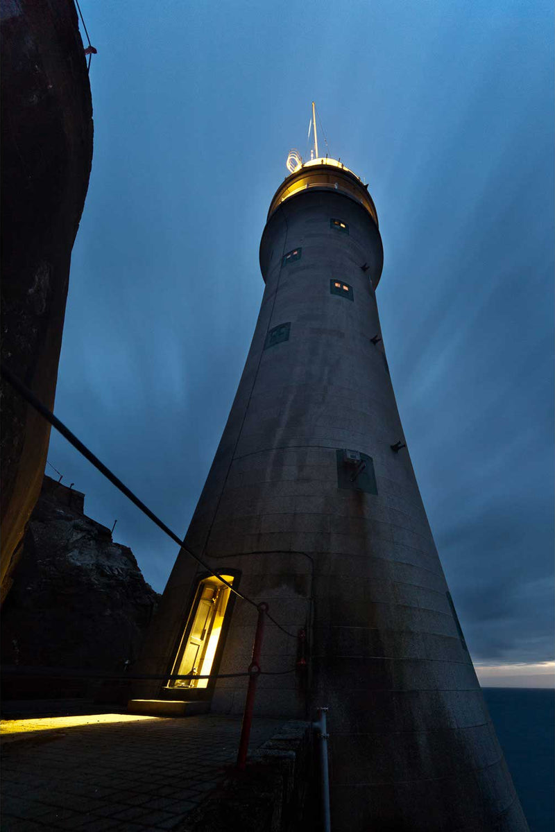 The Fastnet Lighthouse at night, photographed looking straight up the granite tower from its base. The single gunmetal door at the foot of the tower stands open, spilling warm light across the wet paving, with the lantern lit at the top and cloud streaking across the blue sky in a long exposure.