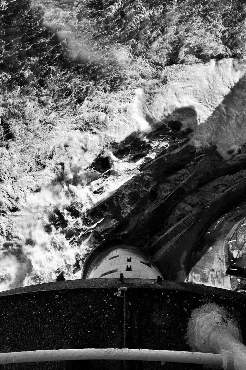 Looking straight down from the balcony of the Fastnet Lighthouse, in black and white. The curved edge of the balcony and its handrail cross the foreground, the granite tower falls away below to the rock at its base with the landing steps and platforms, and heavy Atlantic swell breaks white all around it. Co. Cork. A fine art landscape photograph by Peter Cox.