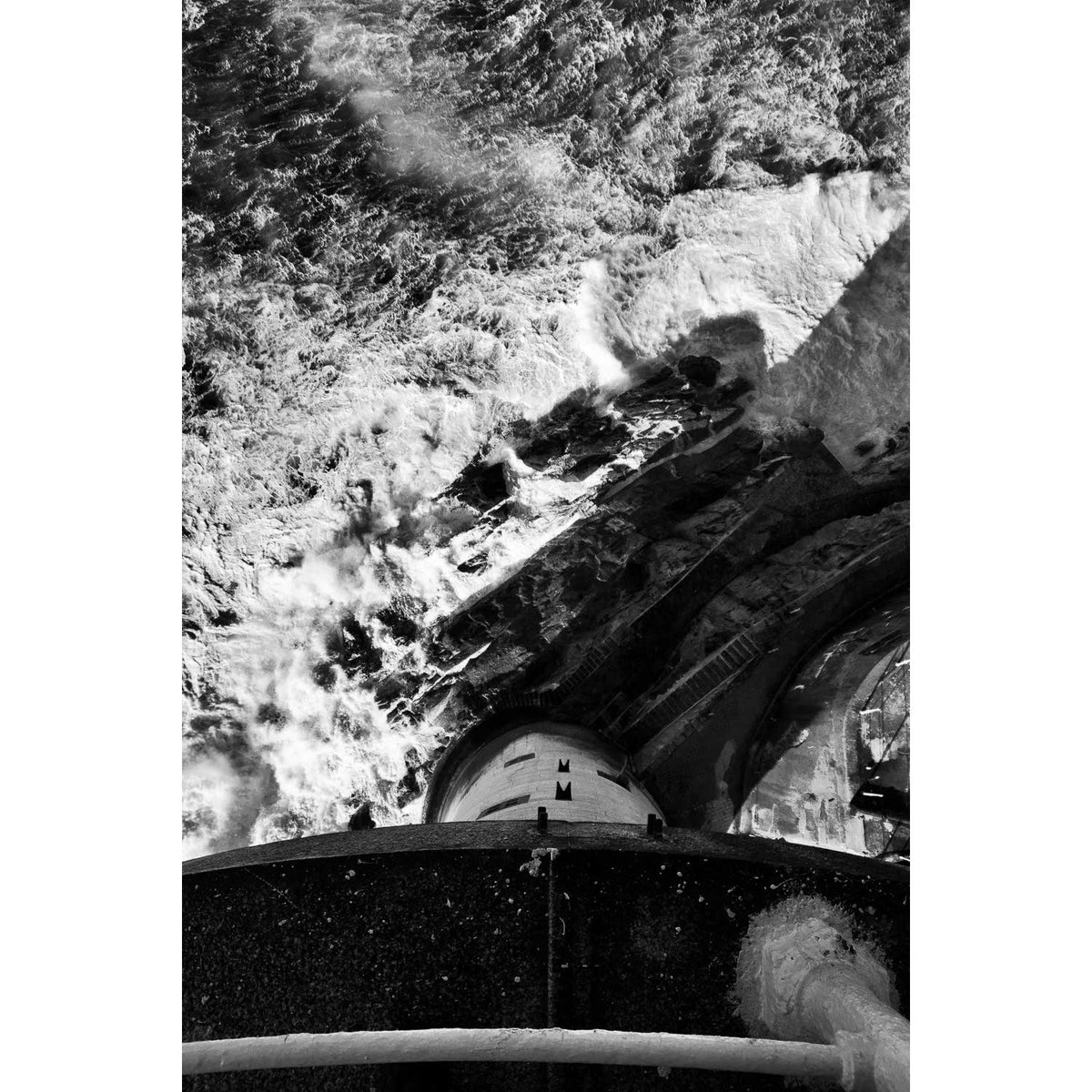 Looking straight down from the balcony of the Fastnet Lighthouse, in black and white. The curved edge of the balcony and its handrail cross the foreground, the granite tower falls away below to the rock at its base with the landing steps and platforms, and heavy Atlantic swell breaks white all around it. Co. Cork. A fine art landscape photograph by Peter Cox.