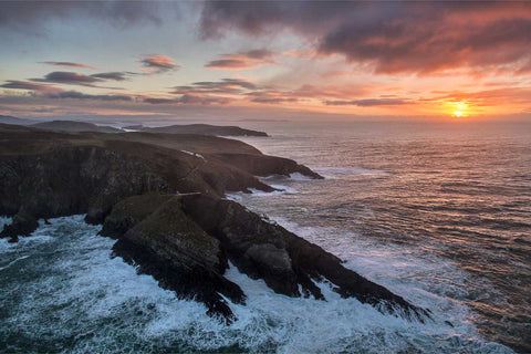 Aerial view of Mizen Head cliffs glowing orange and pink at sunrise with waves crashing below, Cork
