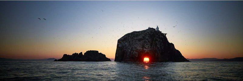 Sunrise at the Bull Rock, off Dursey Island on the Beara Peninsula, Co. Cork. A wide panoramic view of the rock in near silhouette to the centre-right, with the rising sun visible as a small orange disc inside the natural arch at its base, reflected in a broken line across the calm water. The lighthouse and its buildings sit on top of the rock, a lower jagged rock stands to the left, dozens of birds fly across the frame, and the sky graduates from deep blue overhead to orange along the horizon.