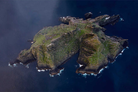 Aerial view looking down on Skellig Michael island with jagged cliffs, green slopes and ancient monastery ruins surrounded by deep blue Atlantic
