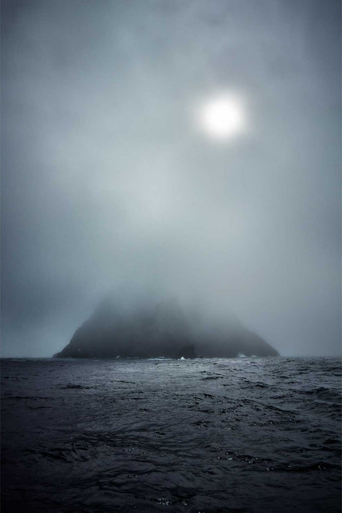 Skellig Michael island emerging as a dark silhouette through thick sea mist with soft sunlight glowing behind, off the Kerry coast