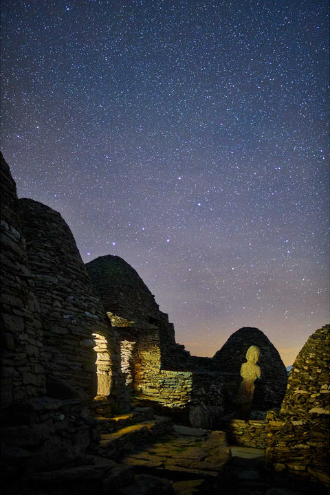 Warm light spilling from beehive huts of the ancient Skellig Michael monastery on a calm night with the sea beyond