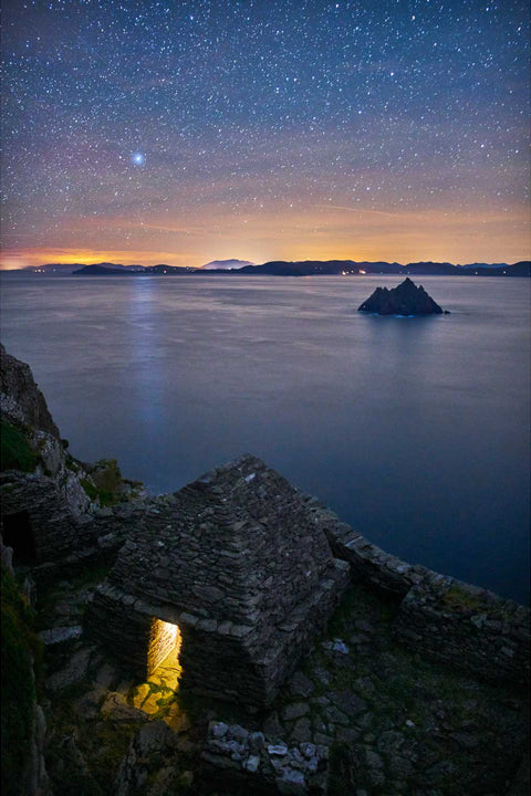 The Small Oratory on Skellig Michael glowing warmly under a starry sky with the Kerry coast and Small Skellig in the background