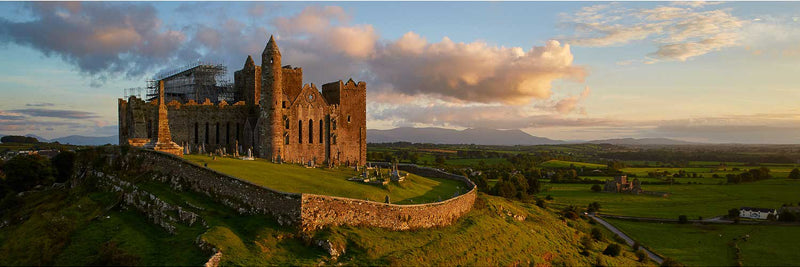 Aerial view of the Rock of Cashel, County Tipperary, at sunset. Medieval cathedral and round tower with conservation scaffolding rise from a limestone outcrop, surrounded by a curving boundary wall and historic graveyard. The Tipperary countryside extends west toward the Galtee Mountains.