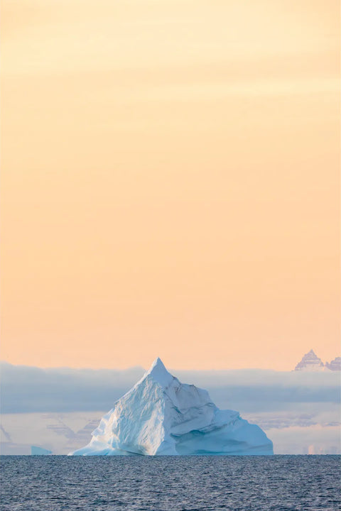 Angular pyramid-shaped iceberg rising from calm water against a warm peach and gold sky in Scoresby Sound, Greenland