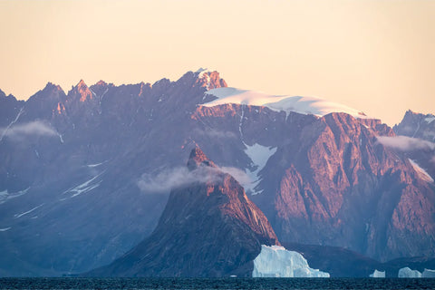 Warm dusk light on enormous mountain peaks and snowfields in Scoresby Sound with an iceberg drifting past, East Greenland