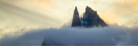 Jagged snow-dusted peaks piercing through low swirling cloud at Arctic dawn in Scoresby Sund, East Greenland, warm amber light on the summits