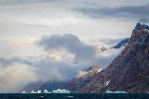 Icebergs drifting beneath cloud-wrapped mountains on dark Arctic waters of Scoresby Sund, Greenland — fine art landscape photograph by Peter Cox
