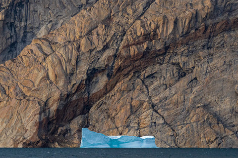 Pale blue iceberg drifting past a towering bronze cliff face in Scoresby Sound, Greenland