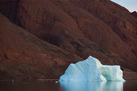Blue-white iceberg floating before rust-red sedimentary cliffs on calm dark water in Scoresby Sound, Greenland