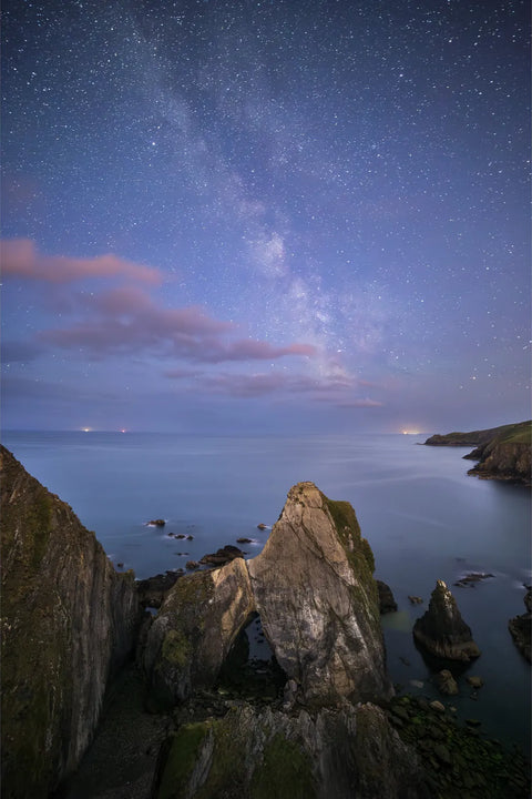 Milky Way stretching across the night sky above Nohoval Cove in Cork, with a limestone sea arch silhouetted against calm Atlantic waters